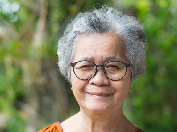 Portrait of a senior woman with short gray hair, wearing glasses, smiling, and looking at the camera while standing in a garden. Space for text. Concept of aged people and healthcare.