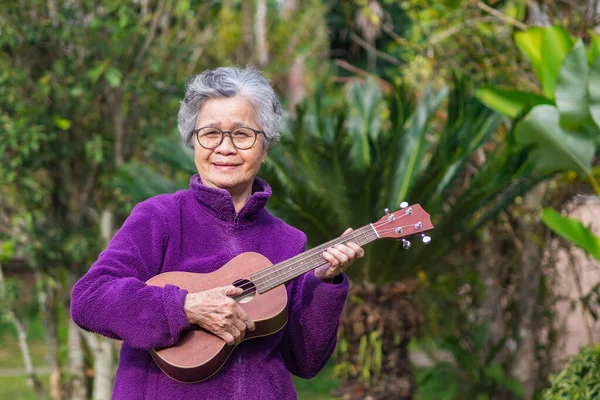Kısa gri saçlı, gözlüklü ve bahçede dikilirken gitar çalan neşeli, yaşlı Asyalı kadın. Yaşlılar ve rahatlama kavramı.
