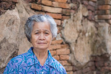 Eski tuğla duvar arka planıyla kameraya bakarken kısa gri saçlı yaşlı Asyalı kadın kameraya bakıyor. Mesaj için yer var. Yaşlılar ve rahatlama kavramı.