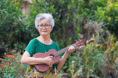 Ukulele çalan, gülümseyen ve bahçede dikilirken kameraya bakan yaşlı bir Asyalı kadının portresi. Mesaj için yer var. Yaşlılar ve rahatlama kavramı.