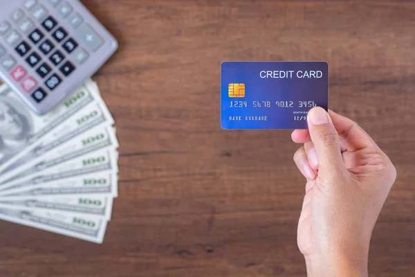 Close-up of hand holding a blue card with US banknotes and a calculator background placed on a wooden table. Space for text. Wood texture background. Business and payment concept.