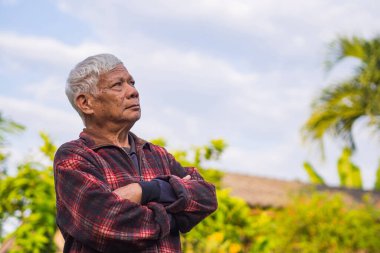Yaşlı Asyalı bir adamın portresi. Gri saçlı, kollarını kavuşturmuş ve bahçede dikilirken yukarı bakıyor. Yaşlılar ve sağlık hizmetleri kavramı.
