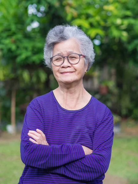 Portrait of an elderly woman with short gray hair,  smiling, arms crossed, and looking at the camera while standing in a garden. Concept of aged people and healthcare.