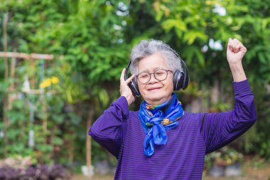 Kısa gri saçlı, gözlüklü, kablosuz kulaklıklı, şarkı dinleyen, bahçede ayakta gülümseyen son sınıf öğrencisi kadın. Yaşlılar ve rahatlama kavramı