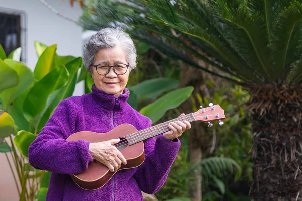 Portrait of an elderly woman playing the ukulele while standing in the garden. Relaxing by singing and play small guitar happy and enjoy life after retiring. Concept of aged people and healthcare.