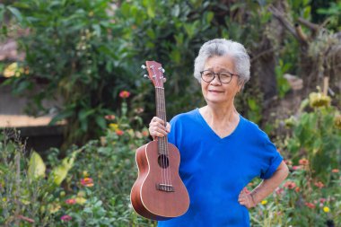 Bahçede dikilirken ukulele tutan son sınıf Asyalı kadın. Şarkı söyleyerek, küçük gitar çalarak, mutlu bir şekilde ve emekli olduktan sonra hayatın tadını çıkararak. Yaşlılar ve sağlık hizmetleri kavramı.