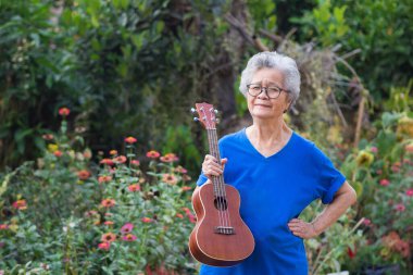 Bahçede dururken elinde ukulele tutan yaşlı bir Asyalı kadının portresi. Şarkı söyleyerek, küçük gitar çalarak, mutlu bir şekilde ve emekli olduktan sonra hayatın tadını çıkararak. Yaşlılar ve sağlık hizmetleri kavramı.
