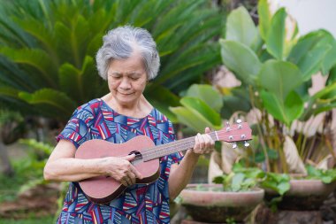 Yaşlı Asyalı kadın bahçede dikilirken ukulele tutuyor ve bakıyor. Şarkı söyleyerek, küçük gitar çalarak, mutlu bir şekilde ve emekli olduktan sonra hayatın tadını çıkararak. Yaşlılar ve sağlık hizmetleri kavramı.