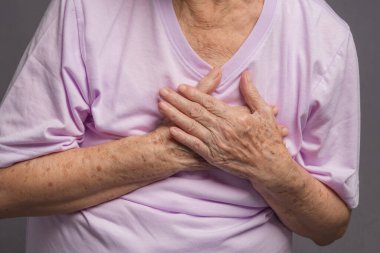 Yaşlı bir kadın gri arka planda dururken göğüs ağrısı çekiyor. Kalp krizi geçirdi. Kardiyoloji hastalığı