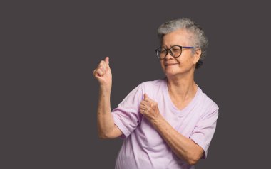 Mutlu yaşlı Asyalı kadın gri bir arka planda dururken gülümseyerek ellerini kaldırıyor ve başka tarafa bakıyor. Yaşlılar ve rahatlama konsepti