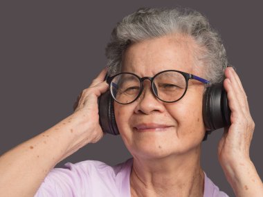 Neşeli, yaşlı, kısa beyaz saçlı, kablosuz kulaklık takan, gri arka planda dururken en sevdiği şarkıyı dinleyen Asyalı bir kadın. Yaşlılar ve rahatlama konsepti