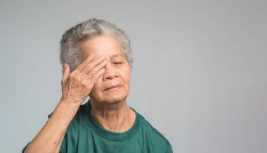 Gri arka planda dururken göz ağrısı çeken yaşlı bir kadının portresi. Yaşlılar ve sağlık hizmetleri kavramı