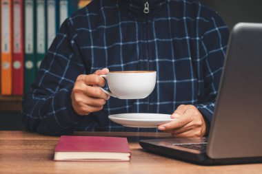 Ofisteki masada oturup çalışırken kazaklı bir iş adamı elinde bir fincan kahve tutuyor. Yakın plan fotoğraf. İçecek ve rahatlama kavramı
