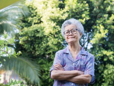 Kısa gri saçlı, güzel, yaşlı Asyalı kadın bahçede kollarını kavuşturmuş dururken gülümseyerek kameraya bakıyor. Mesaj için yer var. Yaşlılar ve rahatlama konsepti