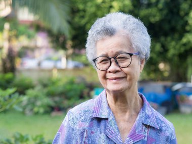 Kısa gri saçlı, güzel, yaşlı Asyalı kadın bahçede dururken kameraya gülümseyerek bakıyor. Mesaj için yer var. Yaşlılar ve rahatlama konsepti