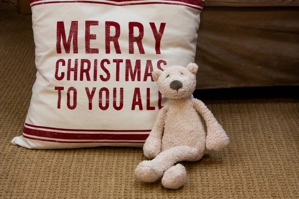 Horizontal Christmas conceptual photo with a white teddy bear toy, sitting home on the carpet near the holiday pillow with printed wishes \