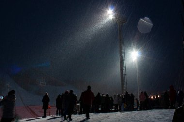 Kışın kar yağarken kayak pistini aydınlatan insan siluetleri ve yüksek lamba direkleriyle kayak merkezinin yatay açık hava fotoğrafı.