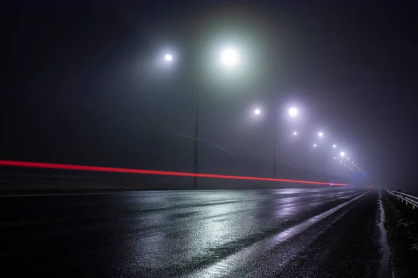 Horizontal conceptual photo of a highway illuinated by street lamps along the road at nigh and red car tail light trail