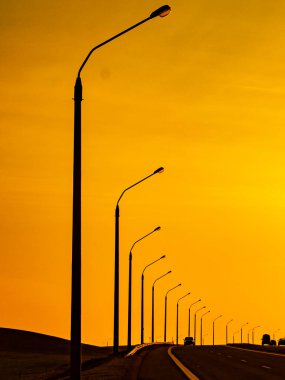 Yol boyunca engelli lamba direklerinin siluetleri ve ufuktaki araçlarla dolu dikey bir açık hava fotoğrafı, sarı akşam gökyüzüne karşı.