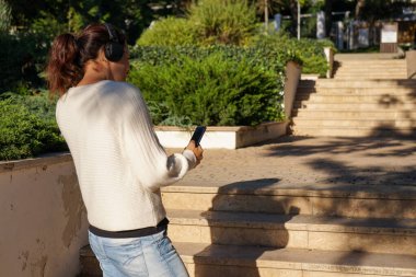 Güz Parkı 'nda yürürken müzik dinlemek için akıllı telefon ve kulaklıklı beyaz örgü kazak ve kot giyen bir kadın.