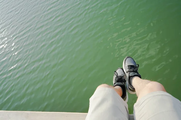 a man dangles his legs on a pier by the lake