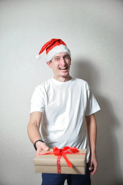 happy man in a white T-shirt and Santa\'s red Christmas hat holds a gift with a red ribbon on hands. congratulations on christmas. holiday concept in pandemic mode. gift delivery to people.