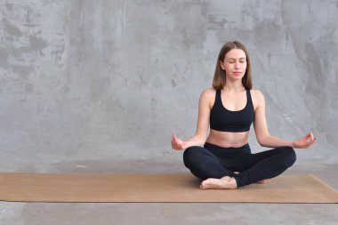 Kahverengi saçlı kadın, gri beton arka planda siyah spor giyerek yoga ve meditasyon pozisyonunda oturuyor. Lotus pozunda meditasyon yapan bir kadın. Rahatlama kavramı. Eğlence ve uyum.