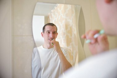 Beyaz tişörtlü bir adam dişlerini banyoda aynanın önünde fırçalıyor. Kişisel Hijyen Konsepti