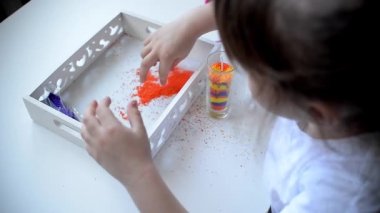 Bir kız küçük parafin taneciklerinden mum yapar. Çocuk kalemleri beyaz masa granüllerinin üzerinde yatıyor. Parafin montessori maddesi üç renkte: sarı, turuncu, mor, makas, mum kabı, fitil.