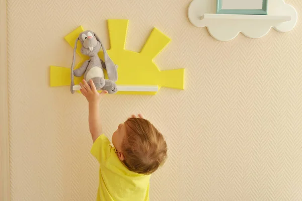 时尚而现代的室内设计 孩子房间的家 蹒跚学步的男孩从架子上拿起一只灰兔的软玩具 — 图库照片