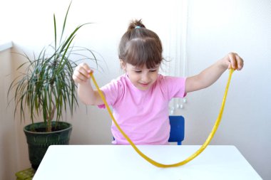 Kız sarı sümüklü sarı sümüğü beyaz bir masada tutuyor ve geriyor. Çocuk balçık oyuncağıyla oynuyor. Sümük yapıyorum. Uzayı kopyala Diy konsepti. Deney bilimsel yöntemi zamanı..