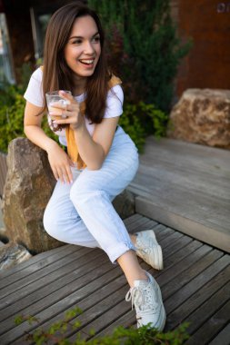 Çekici, özgür, sıradan görünümlü genç bir kadın sokakta kahve içiyor.