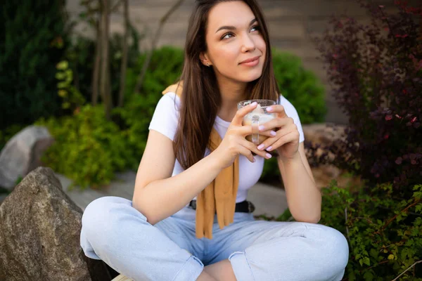 Šťastná mladá žena sedí v krásném parku a pije pikantní latté — Stock fotografie