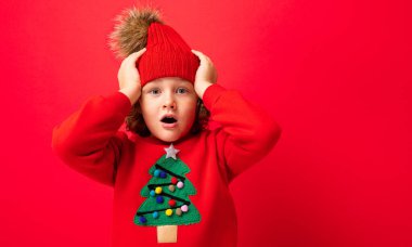Kırmızı arka planda Noel ağacı olan, kalın şapkalı ve süveterli havalı sarışın çocuk etrafta dolanıyor, Noel konsepti.