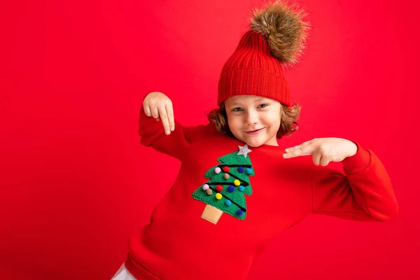 Adolescent cool dans un pull de Noël rouge s'amuser autour sur le fond d'un mur rouge, un chapeau chaud et un pull avec un arbre de Noël — Photo