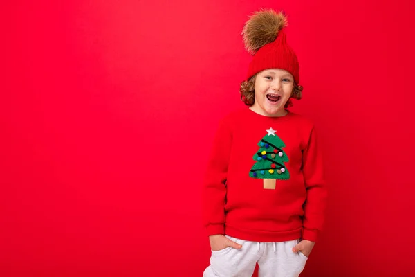 Modelo de niño sobre un fondo rojo, retrato de una rubia guay con rizos en un suéter con un árbol de Navidad — Foto de Stock