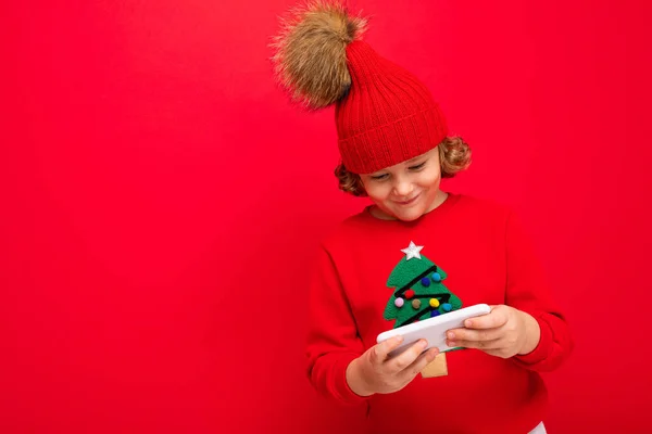 Un jeune homme avec un smartphone dans les mains sur le fond d'un mur rouge, dans un chapeau tricoté et un pull avec un arbre de Noël. — Photo