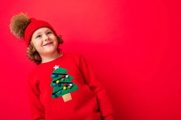 Lindo chico rubio en sombrero caliente y suéter de Navidad sobre fondo rojo con sonrisa en su cara — Foto de Stock