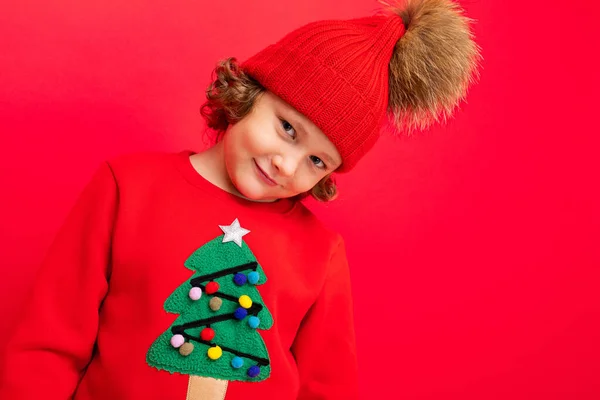 Chico fresco con rizos sobre un fondo rojo en un suéter con un árbol de Navidad — Foto de Stock