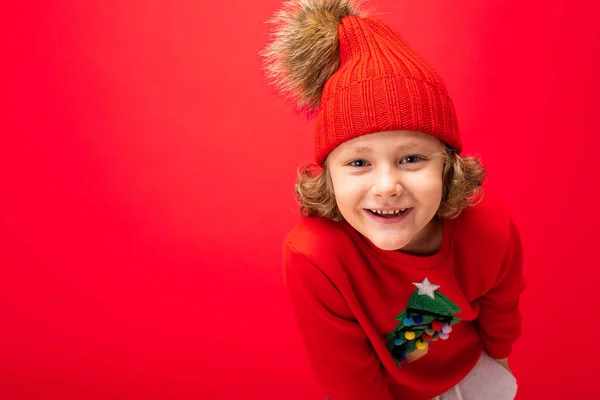 따뜻 한 모자를 쓰고 크리스마스 트리가 달린 스웨터를 입은 멋진 금발의 아이가 빨간 배경 위에서 크리스마스 컨셉트로 장난치고 있습니다. — 스톡 사진