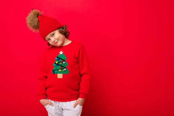 Δροσερό αγόρι με μπούκλες σε κόκκινο φόντο σε ένα πουλόβερ με ένα χριστουγεννιάτικο δέντρο — Φωτογραφία Αρχείου