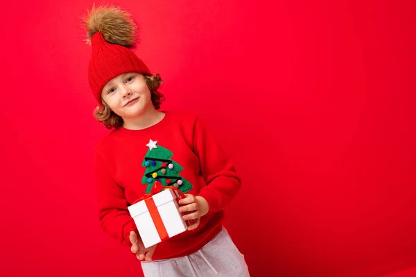 En cool pojke med lockar på en röd vägg bakgrund i en tröja med en julgran håller en presentask i händerna — Stockfoto