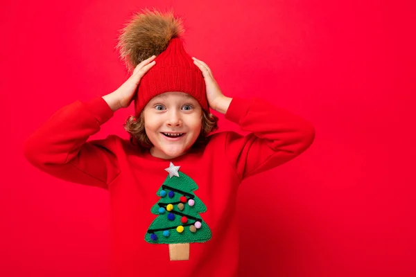 Söt blond pojke i varm hatt och jul tröja på röd bakgrund med leende på ansiktet — Stockfoto