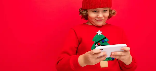 En ung man med en smartphone i händerna mot bakgrund av en röd vägg, i en stickad hatt och en tröja med en julgran. — Stockfoto