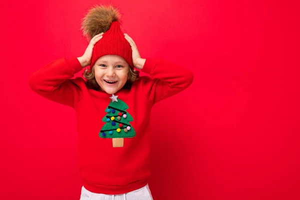 Niño rubio fresco en sombrero caliente y suéter con árbol de Navidad sobre fondo rojo tonteando alrededor, concepto de Navidad — Foto de Stock
