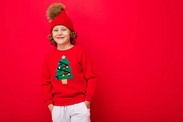 Cool blond enfant en chapeau chaud et pull avec arbre de Noël sur fond rouge folie autour, concept de Noël — Photo