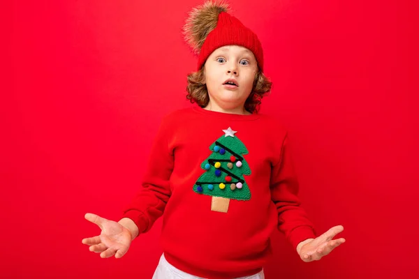 Pojke modell på en röd bakgrund, porträtt av en cool blondin med lockar i en tröja med en julgran — Stockfoto