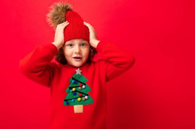 Kırmızı arka planda, sıcak şapkası ve süveteri olan, Noel ağacı olan neşeli, komik çocuk.