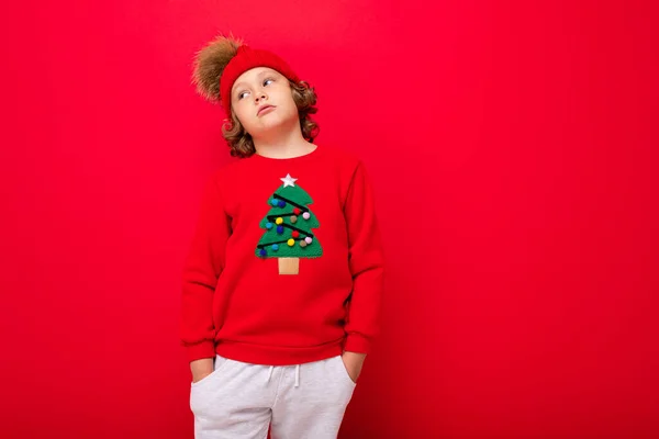 Cool blond enfant en chapeau chaud et pull avec arbre de Noël sur fond rouge folie autour, concept de Noël — Photo