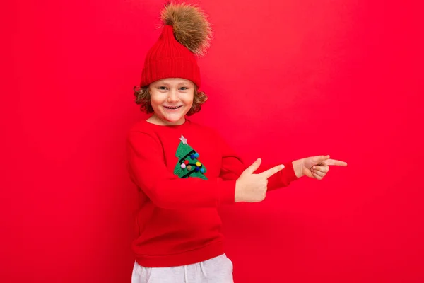 Adolescente fresco en un suéter rojo de Navidad jugando alrededor contra el fondo de una pared roja, un sombrero caliente y un suéter con un árbol de Navidad — Foto de Stock